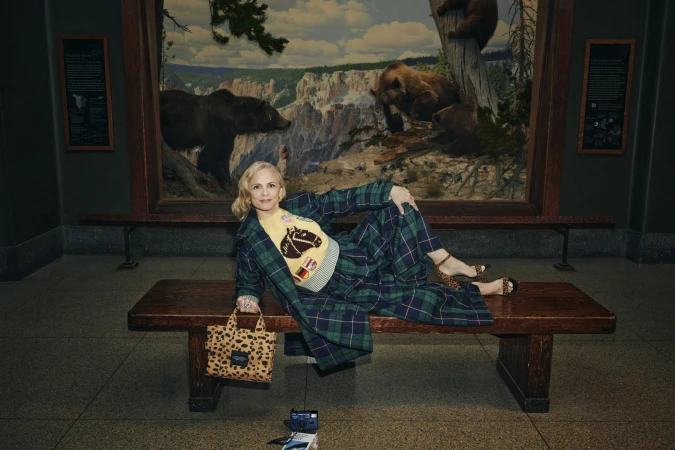 Woman in plaid coat and matching trousers lying on a wooden bench inside a museum with a bear diorama background.