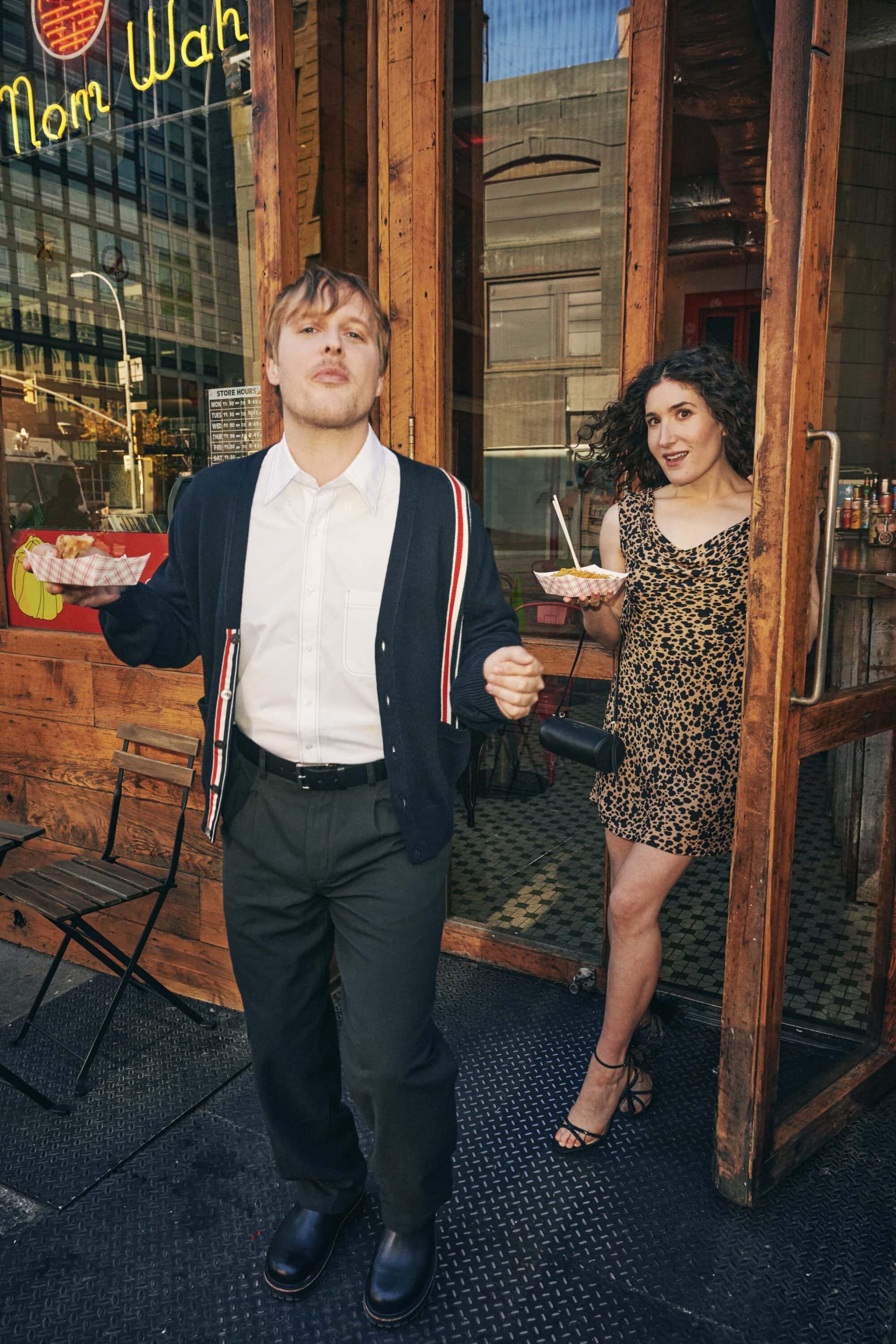 Two models standing outside a restaurant entrance holding food trays, featuring a man in a dark cardigan and a woman in a leopard dress.
