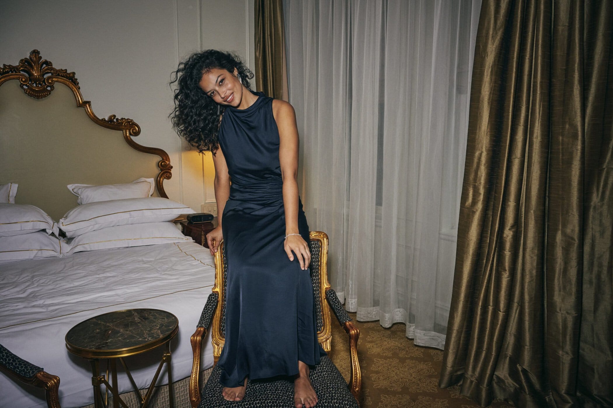 Woman with curly dark hair wearing a navy blue satin dress seated on a gold-trimmed armchair in a hotel room.
