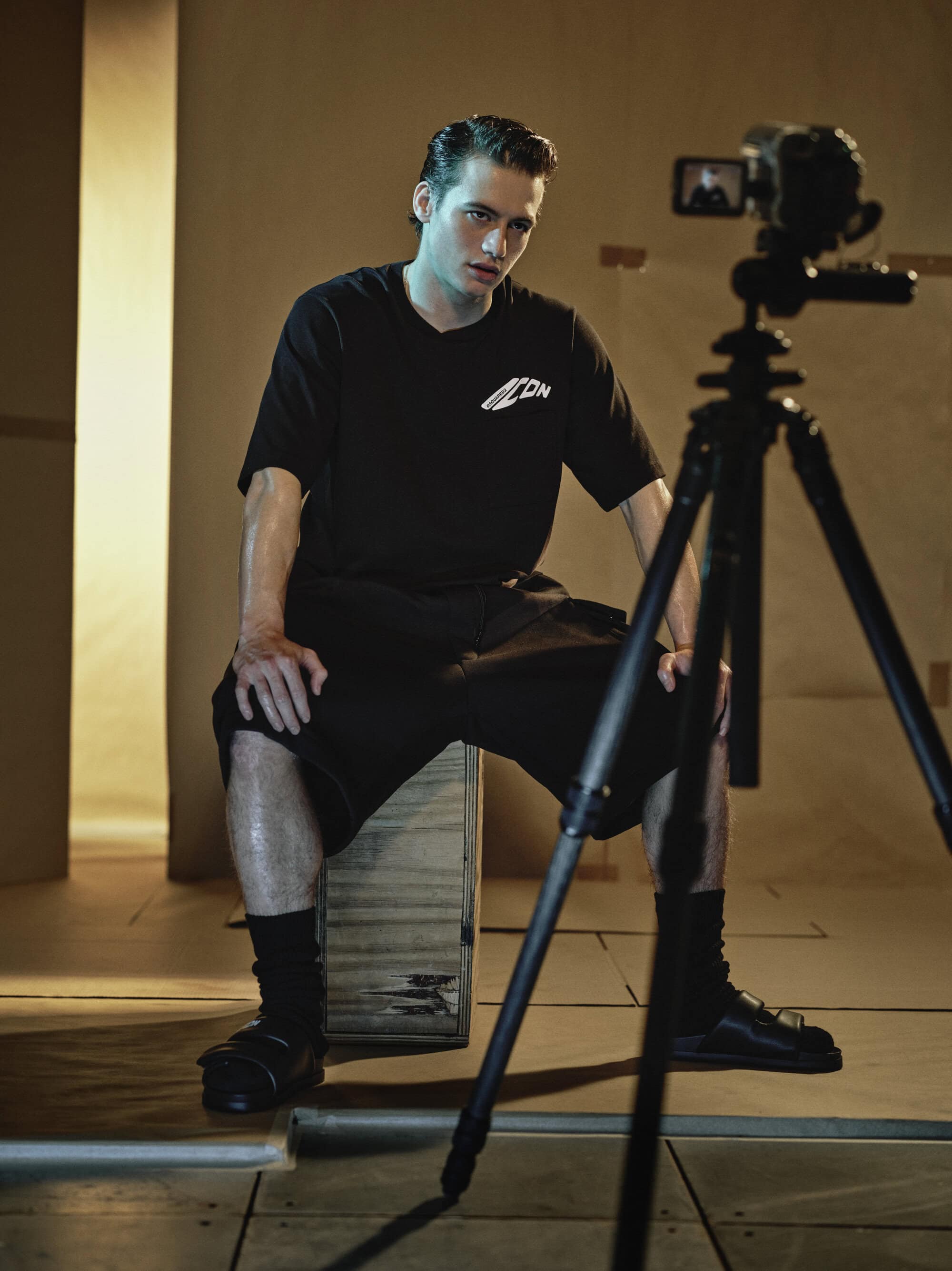 Model wearing black t-shirt and shorts seated on a crate with camera tripod in foreground