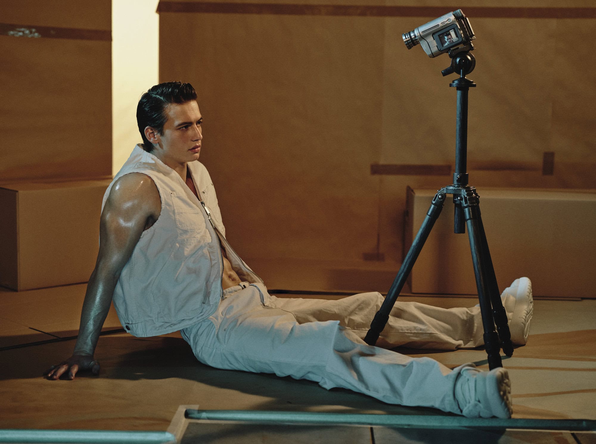 A young male model seated on the floor in white pants and vest, looking towards a vintage camera on a tripod.