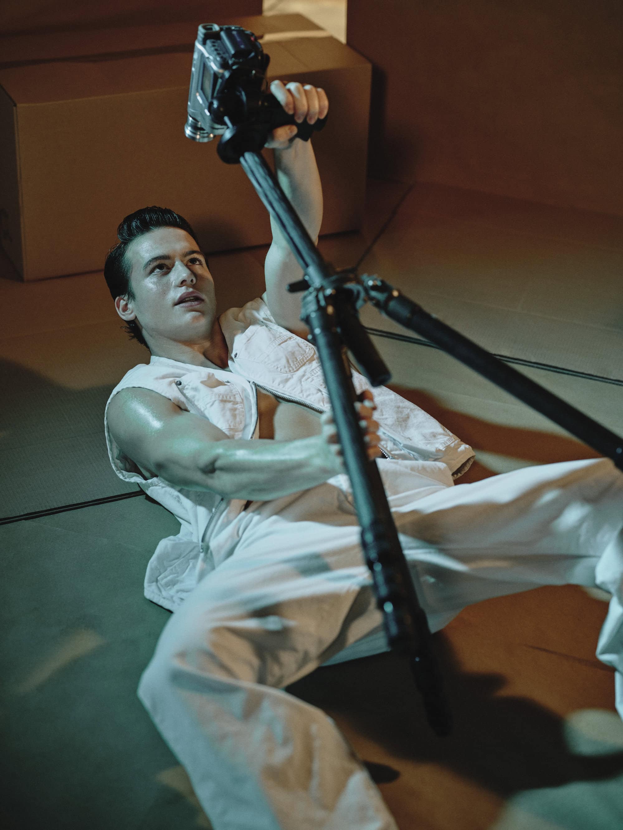 Man wearing white vest and trousers lying on floor holding a camera tripod in Dsquared2 campaign