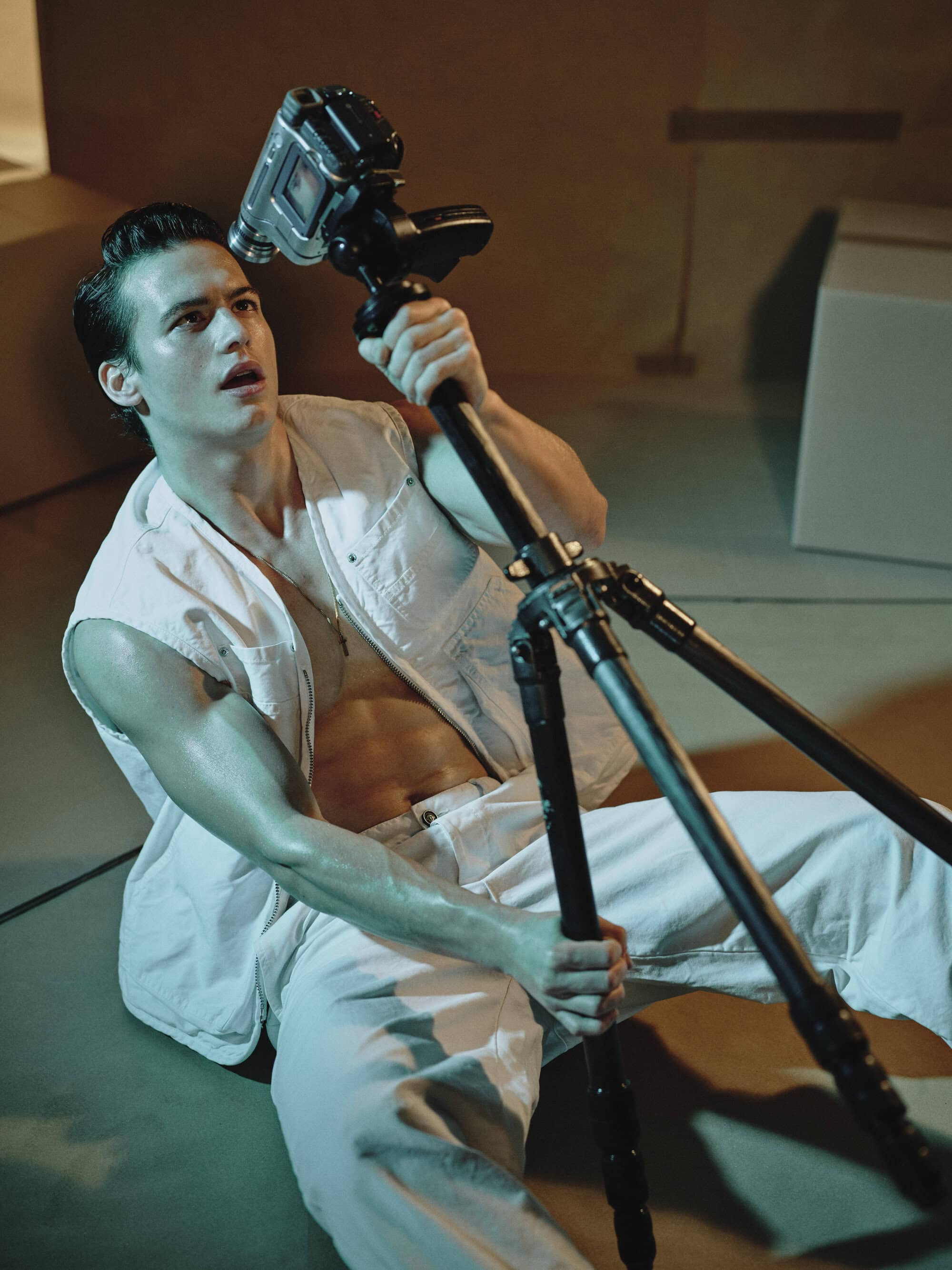 Man wearing a white sleeveless vest and trousers holding a camera tripod in an indoor setting