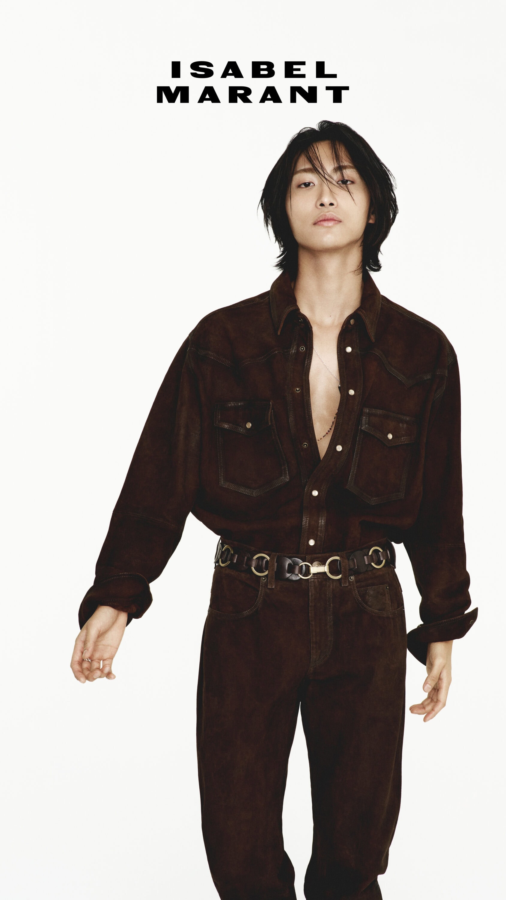 Man with dark hair wearing brown button-down shirt and matching trousers against a solid white background