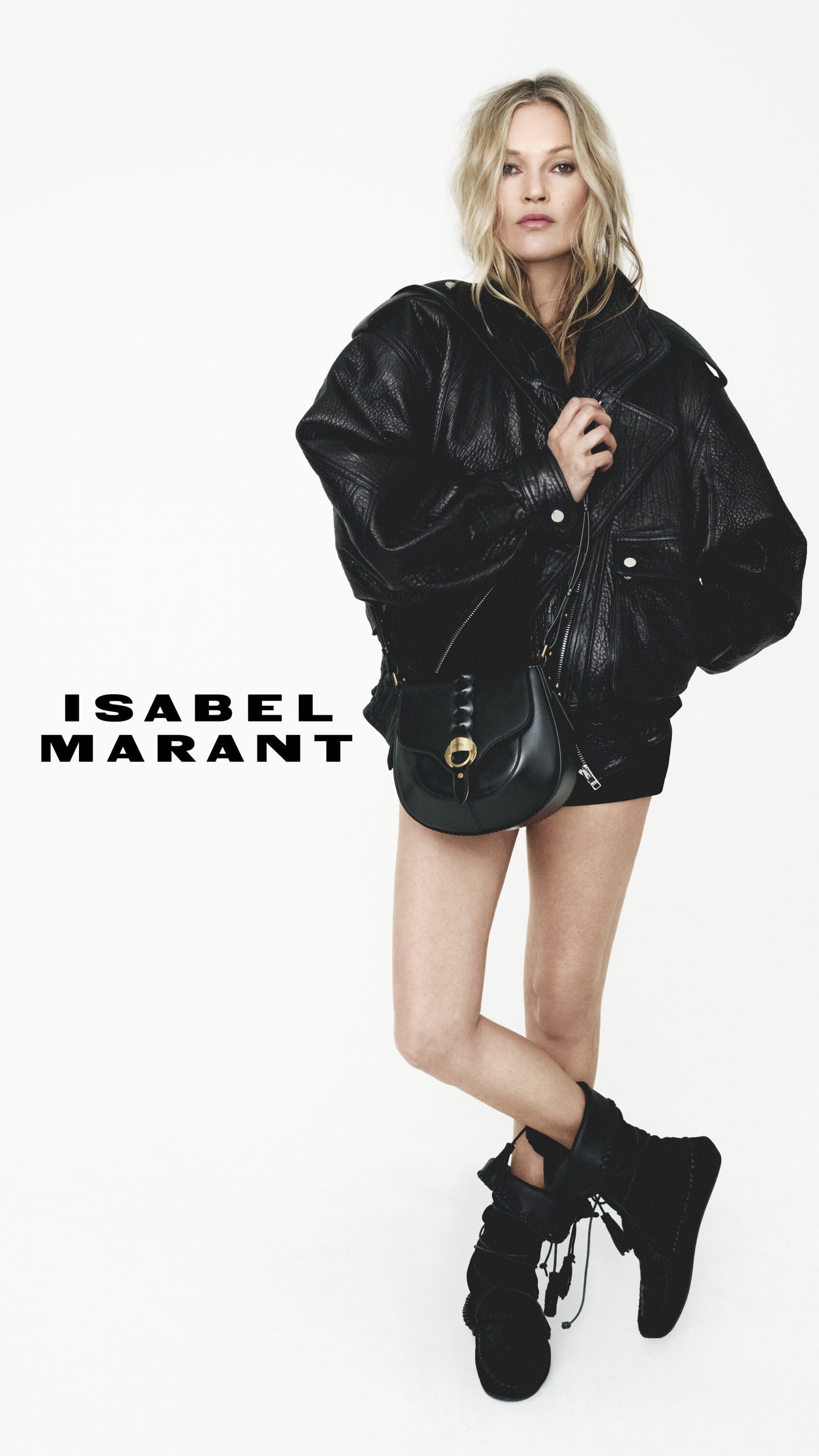 Kate Moss wearing an oversized black leather jacket and black shoulder bag against a plain white background