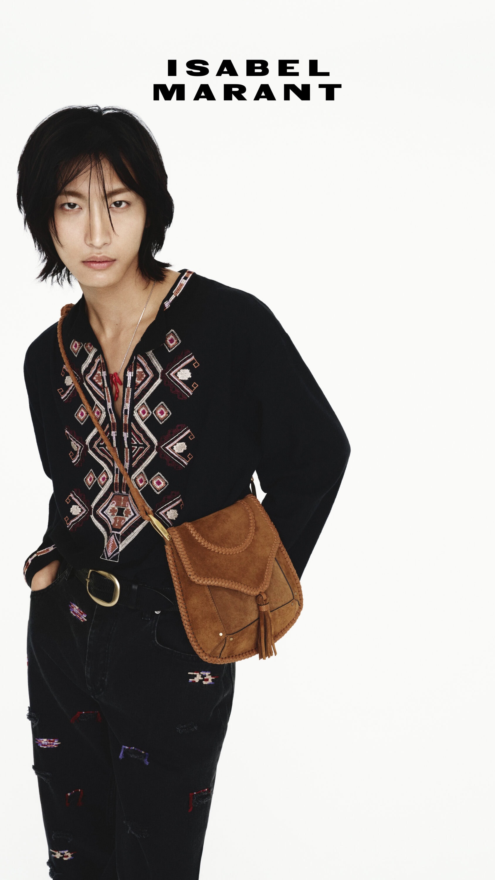 Man with black hair wearing embroidered black shirt and brown leather crossbody bag against white background