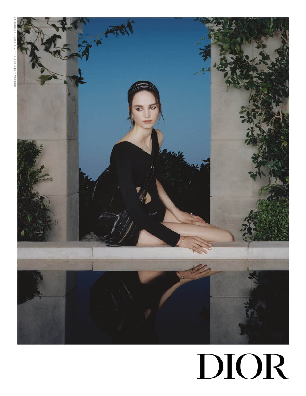 Female model wearing a black top and headband seated on a ledge with a reflection below, framed by architectural stone pillars.