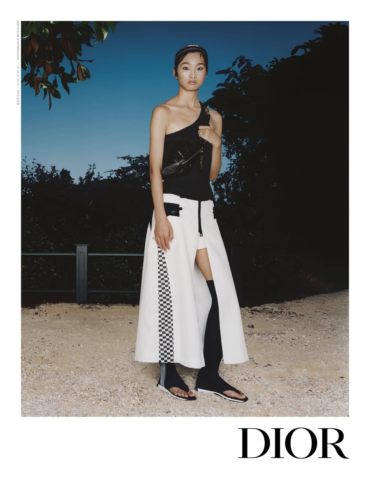 Model standing outdoors at dusk wearing a black one-shoulder top and white split skirt with checkered detailing