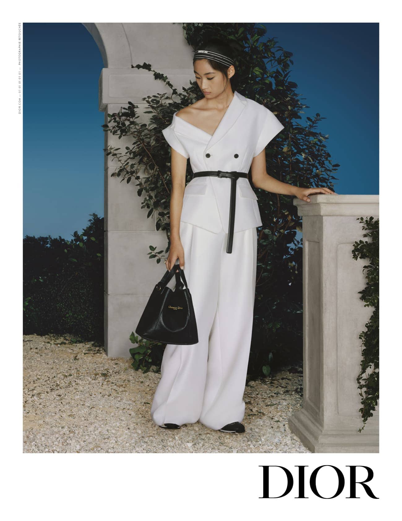 Model in a white Dior outfit with a black belt and handbag standing outdoors near stone architecture