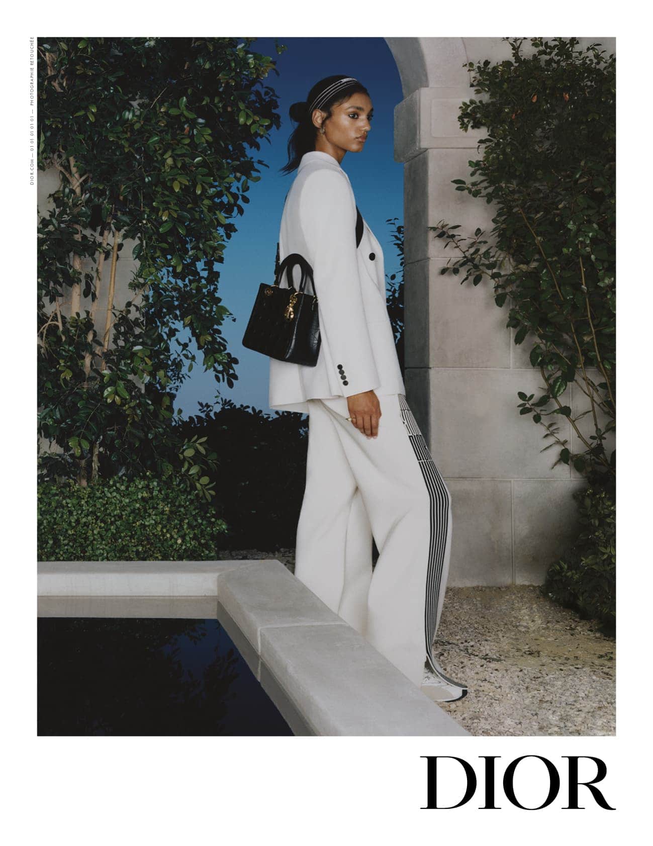 Model in white suit and black handbag standing outdoors near a stone archway and reflecting pool for Dior campaign