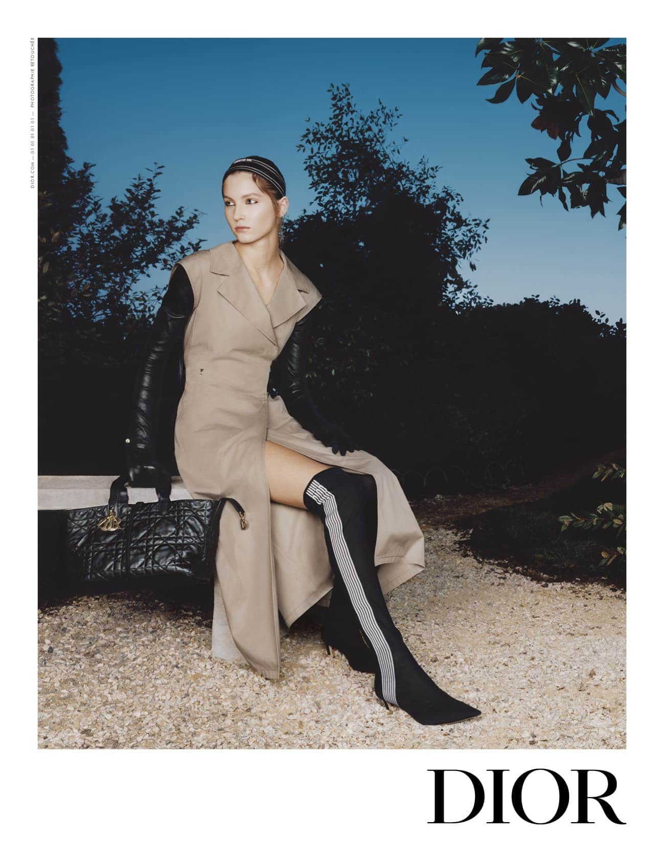 Model wearing a tan sleeveless coat and black boots seated outdoors holding a black quilted handbag against twilight sky