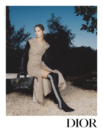 Model wearing a tan sleeveless coat and black boots seated outdoors holding a black quilted handbag against twilight sky