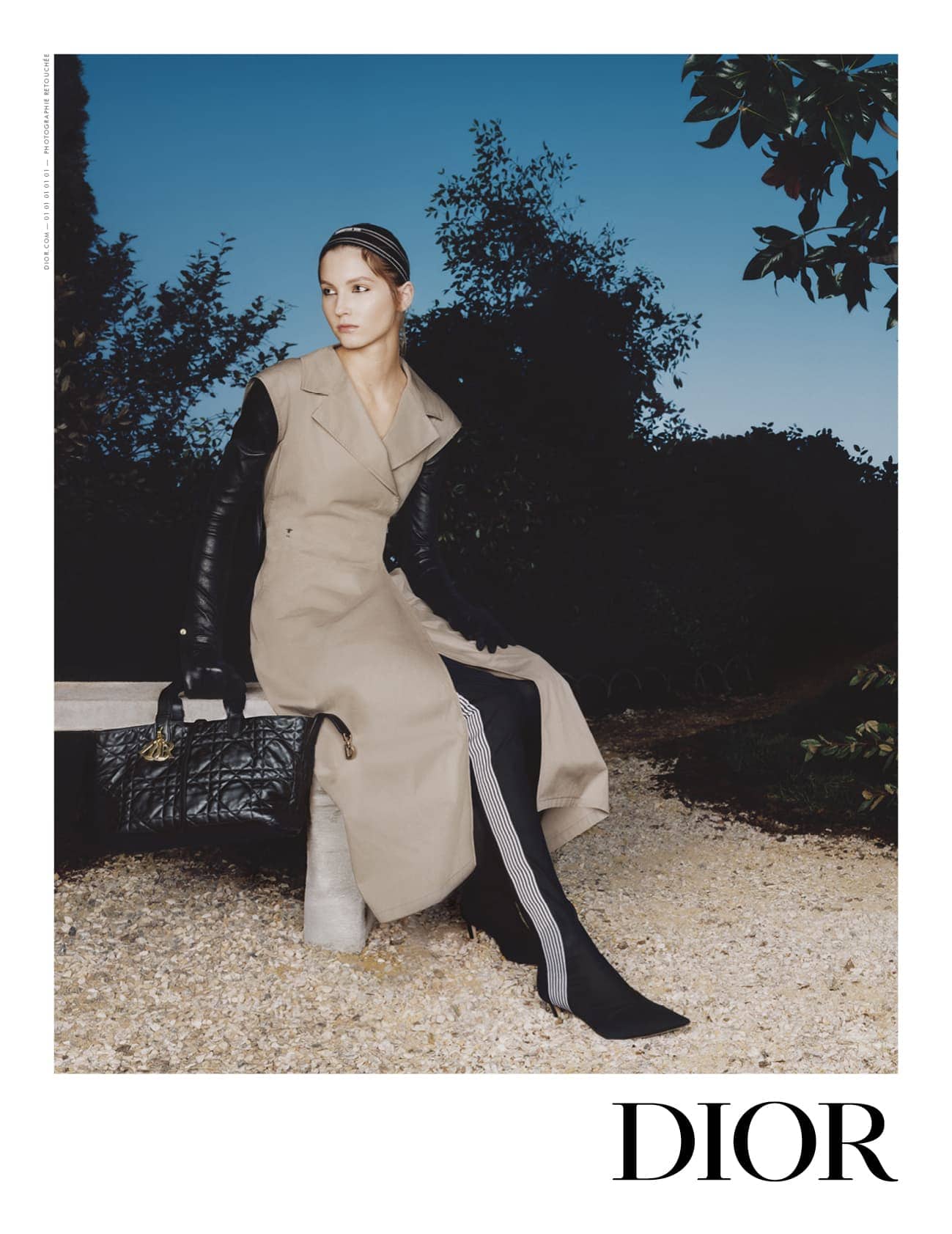 Model in a tan coat and black gloves seated on a stone bench in a garden under a twilight sky, holding a quilted black handbag.
