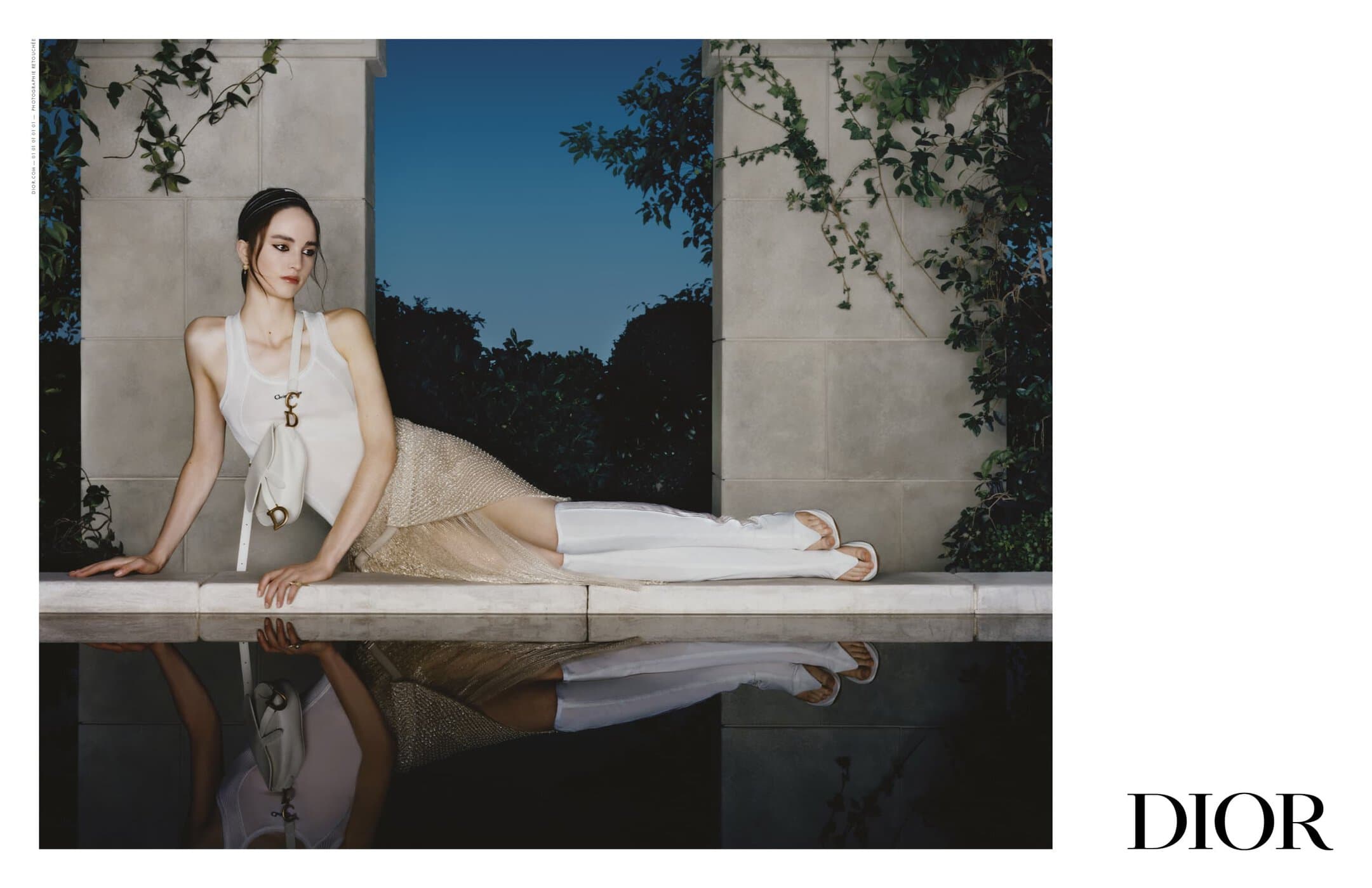 Model reclining by a reflecting pool wearing a white tank top and skirt with white boots for Dior Spring 2025 campaign.