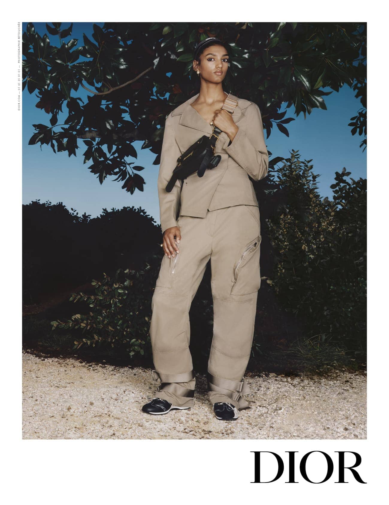 Model standing outdoors wearing a tan jacket and trousers holding a black crossbody bag under a tree.