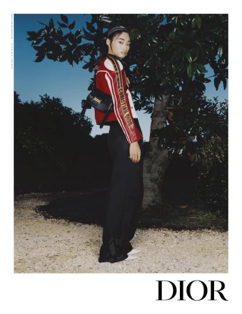 Model standing outdoors near a tree at dusk wearing a red and white jacket with dark trousers for Dior Spring 2025 campaign.