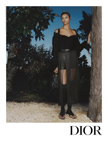 Model standing outdoors near a tree in a dark off-shoulder top and fringe skirt, holding a small handbag