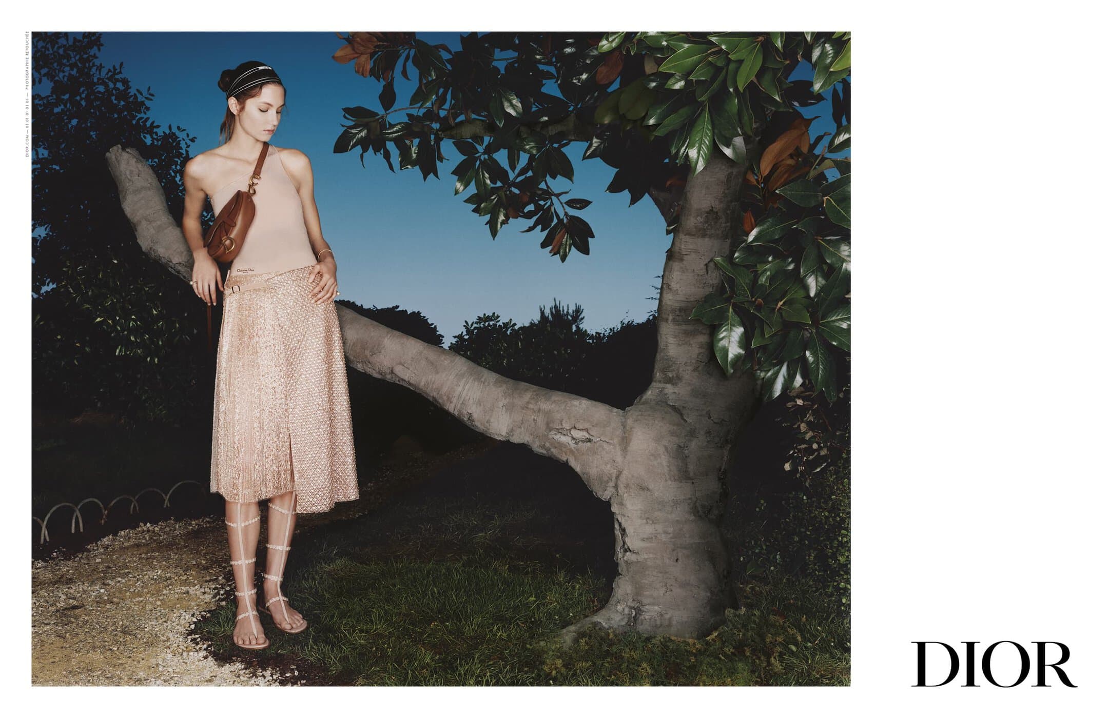 Model leaning against a large tree branch wearing a cream top and patterned skirt with gladiator sandals for Dior