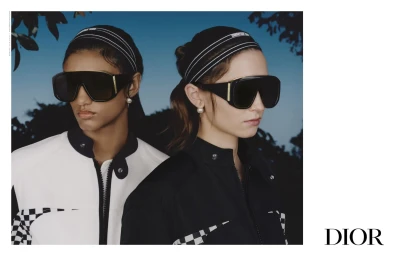 Two female models wearing black oversized sunglasses and branded headbands against an outdoor sky backdrop for Dior