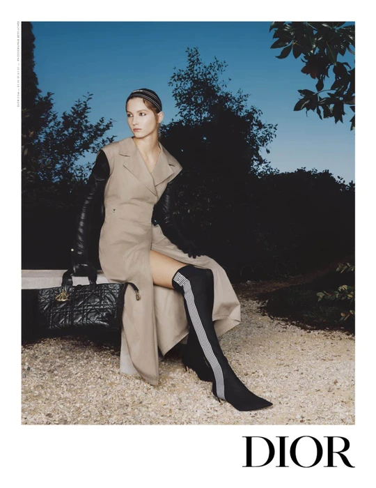Model wearing a tan sleeveless coat and black boots seated outdoors holding a black quilted handbag against twilight sky