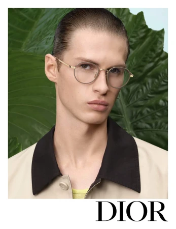 Portrait of a young male model wearing wire-rimmed glasses and a collared jacket against a background of large green tropical leaves.