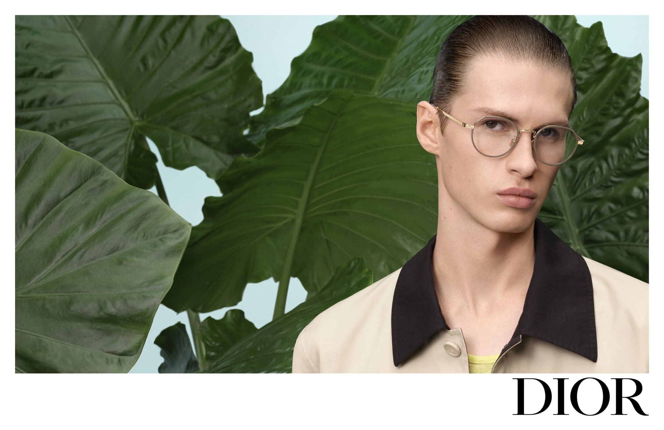 Model wearing thin-rimmed glasses and a beige jacket against large green tropical leaves for Dior Spring 2025 ad campaign.