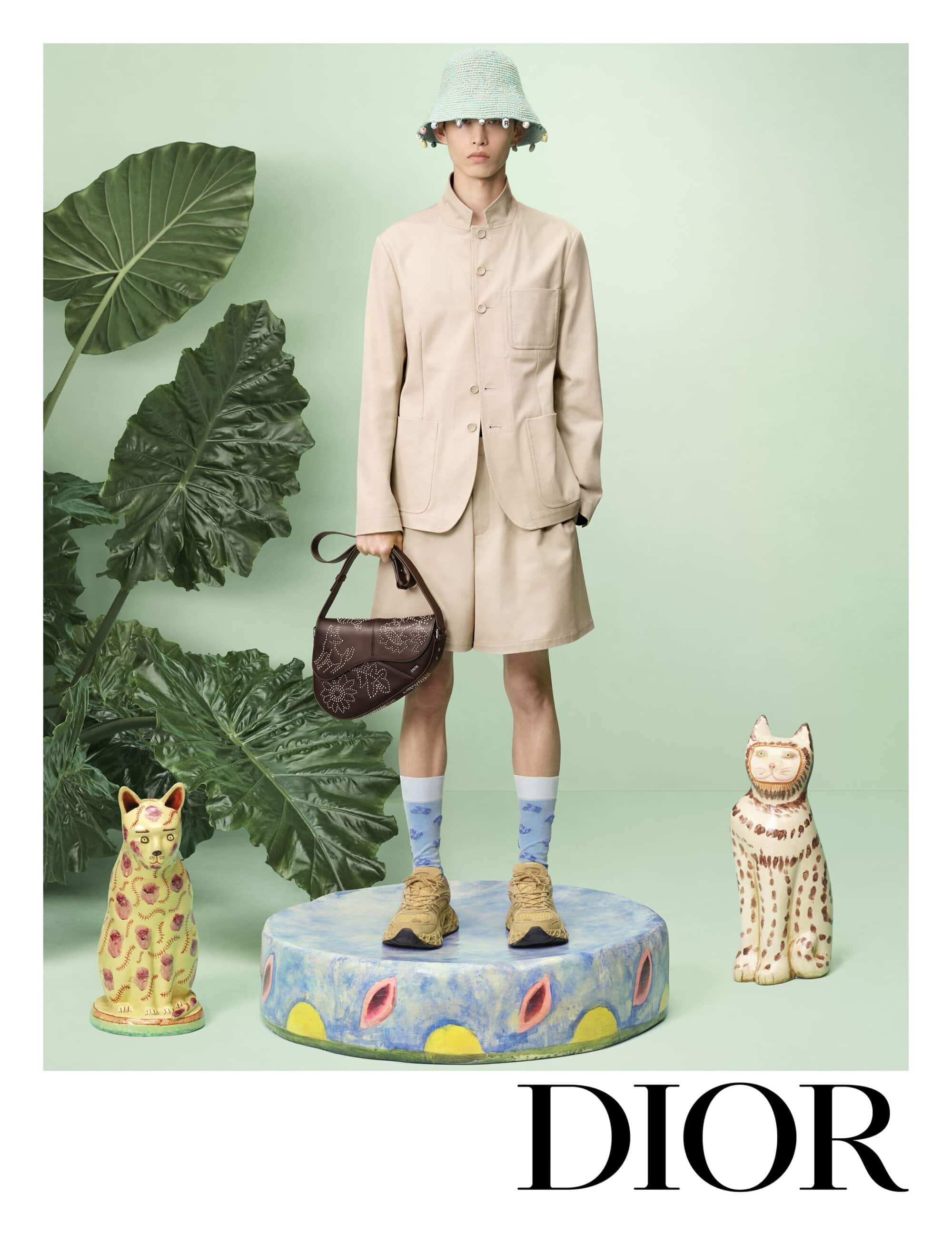 Model standing on a decorated round plinth wearing a beige jacket, shorts, hat, and holding a brown handbag against a pale green backdrop.