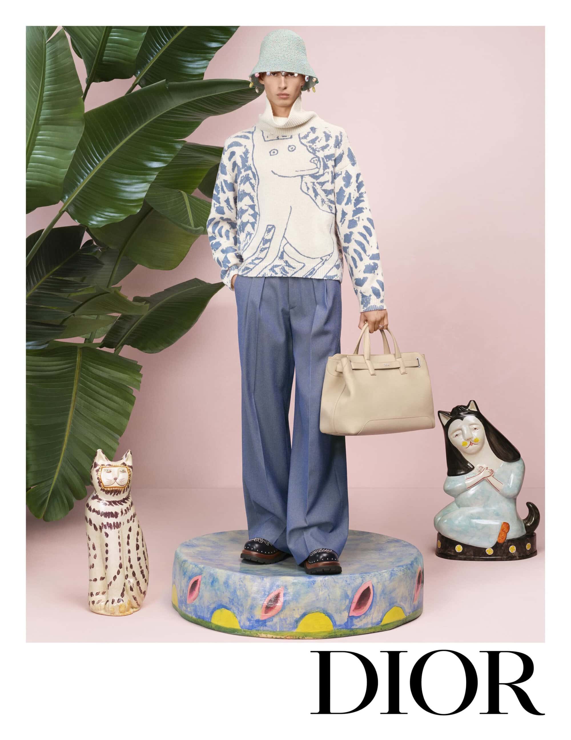 Model wearing a bucket hat and patterned sweater holding a handbag while standing on a painted platform with ceramic cats.