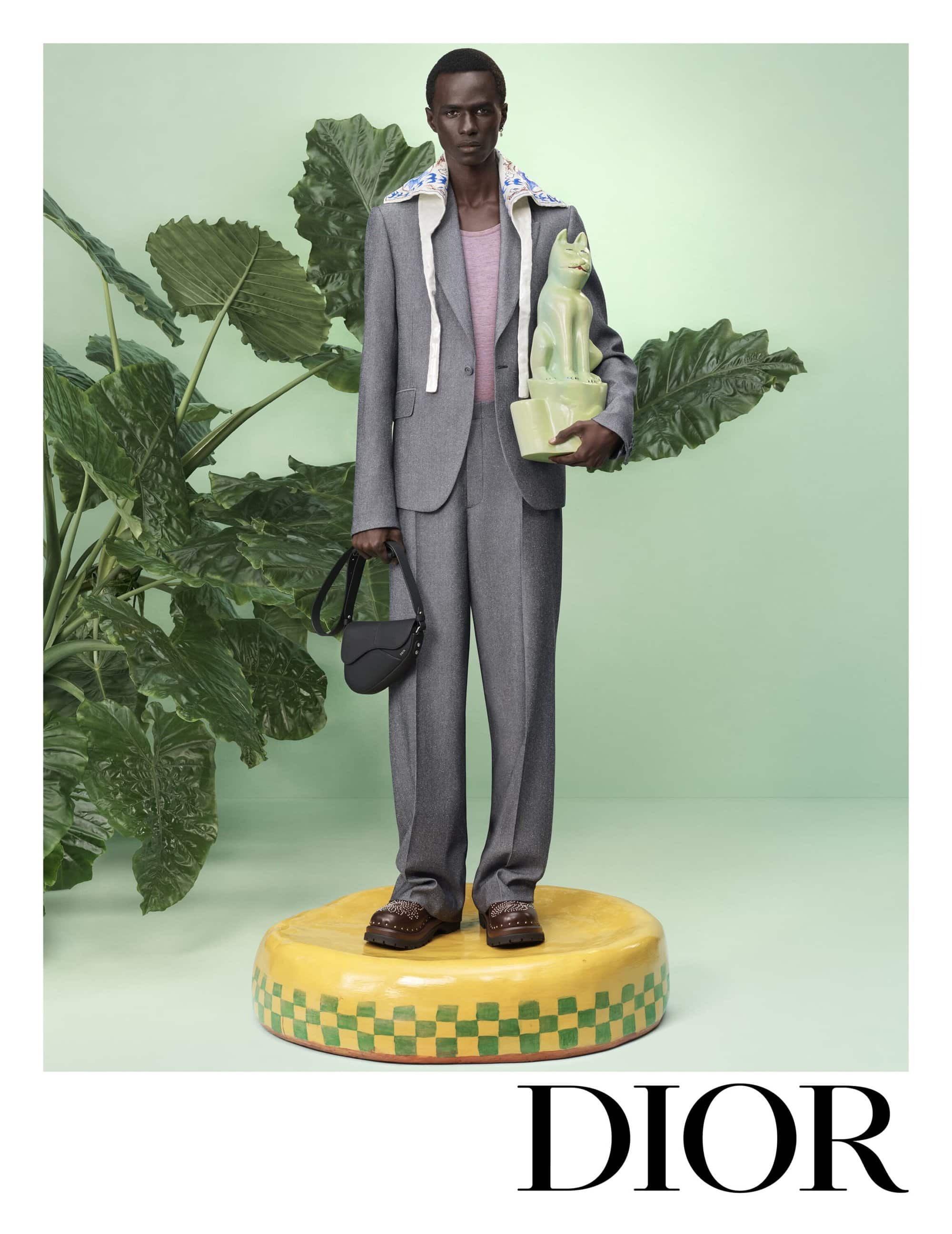 Model in a grey suit holding a ceramic sculpture and a black saddle bag standing on a yellow platform