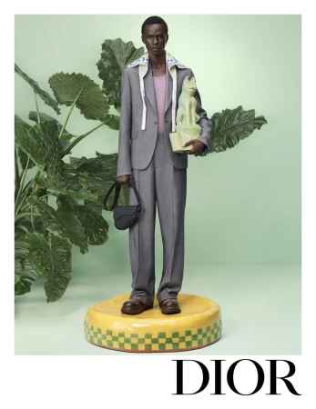 Model in a grey suit holding a ceramic sculpture and a black saddle bag standing on a yellow platform