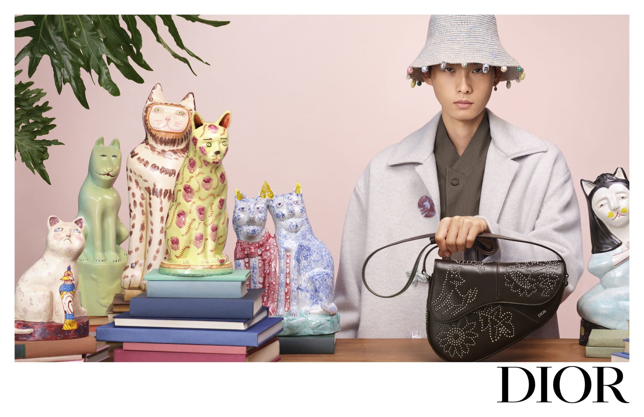 Model in a pale coat and beaded bucket hat holding a dark leather saddle bag amidst colourful ceramic cat figurines