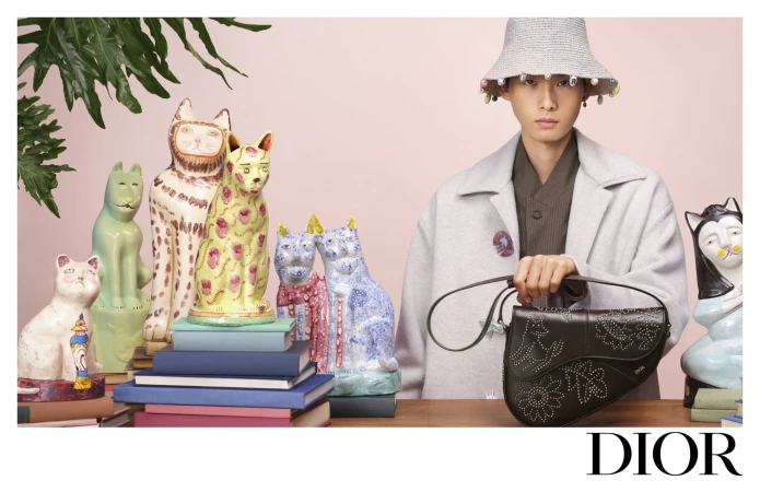 Model in a pale coat and beaded bucket hat holding a dark leather saddle bag amidst colourful ceramic cat figurines