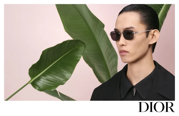 Close-up portrait of a male model wearing dark sunglasses and a black collared shirt against a pale backdrop with large green leaves
