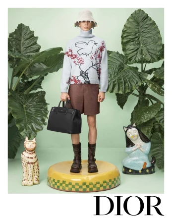 Model standing on a yellow pedestal holding a black handbag wearing a turtleneck sweater and bucket hat amidst large tropical leaves