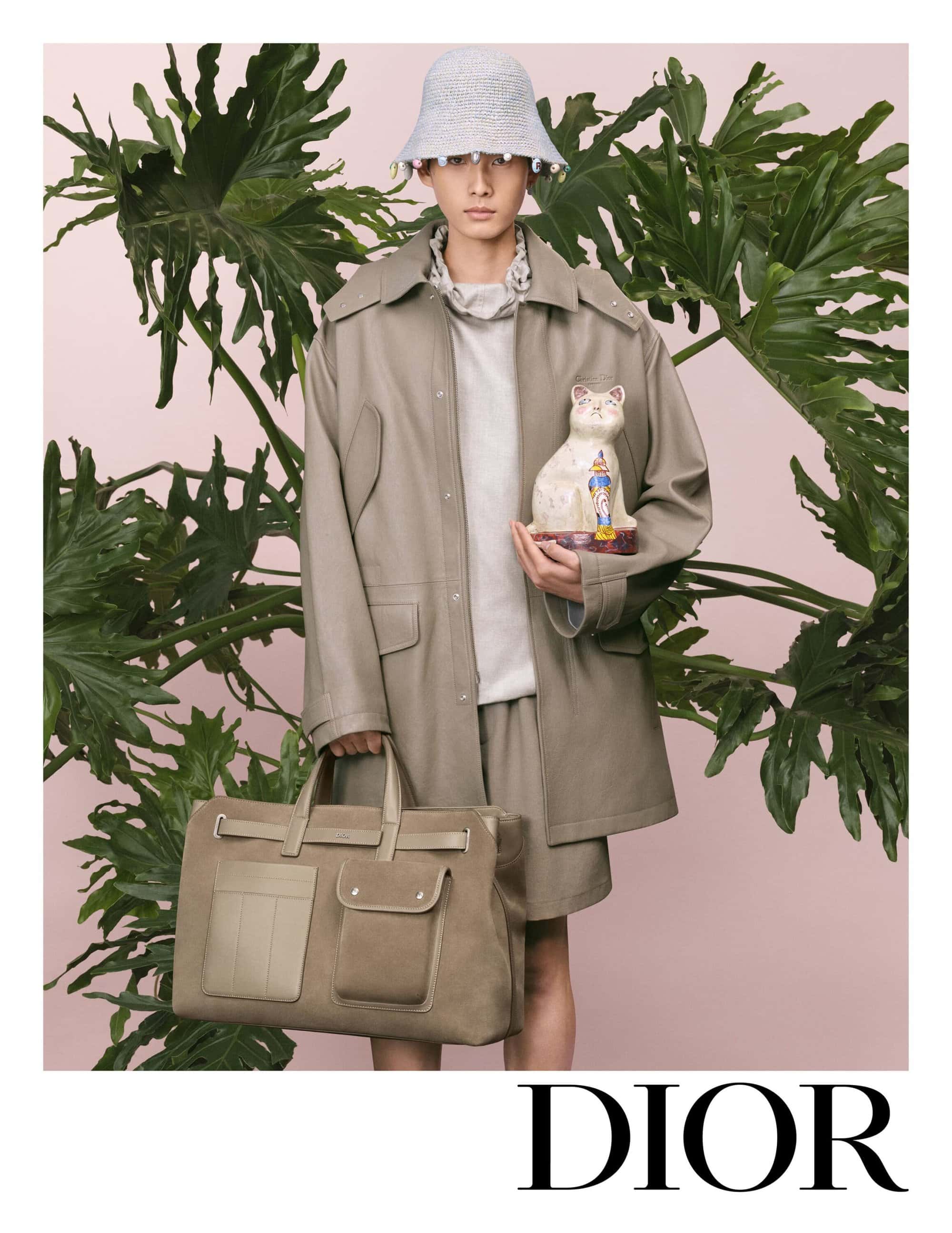 Model wearing a bucket hat, oversized jacket, and shorts while holding a ceramic cat and a large duffel bag before large tropical leaves.