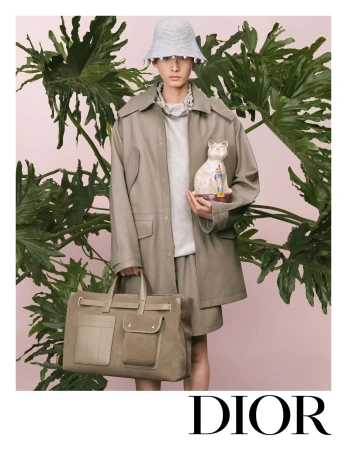 Model wearing a bucket hat, oversized jacket, and shorts while holding a ceramic cat and a large duffel bag before large tropical leaves.