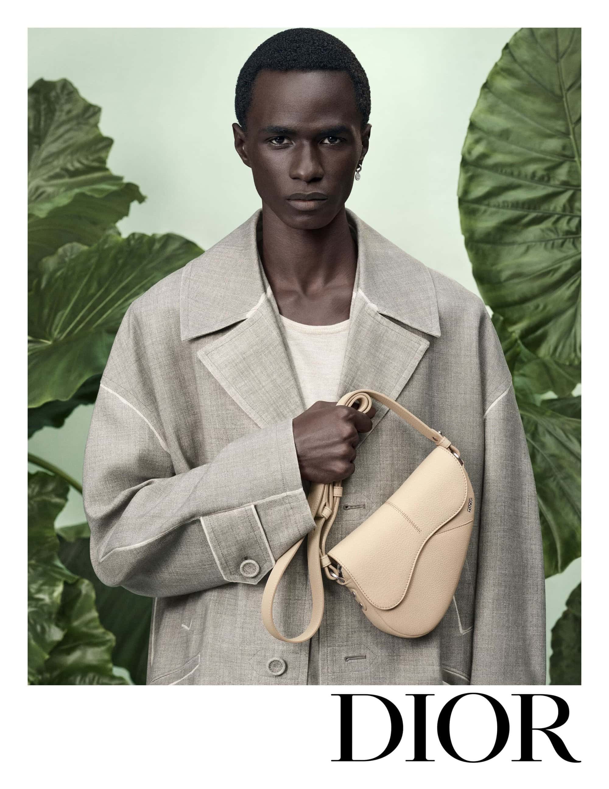 Black man wearing grey coat holding a beige saddle bag against tropical green leaves