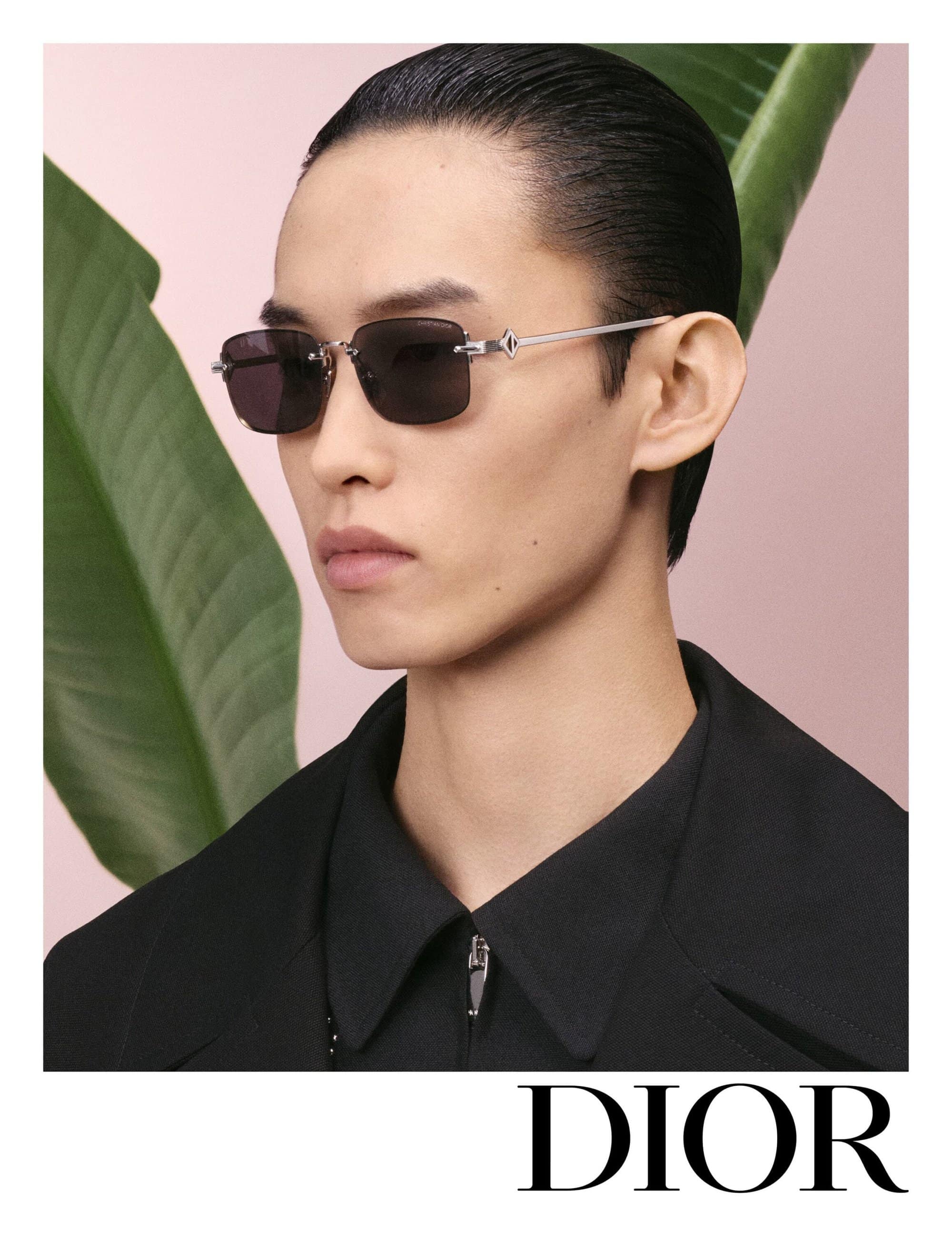 Close-up portrait of a man wearing dark sunglasses and a black shirt against a pink wall with large green leaves.