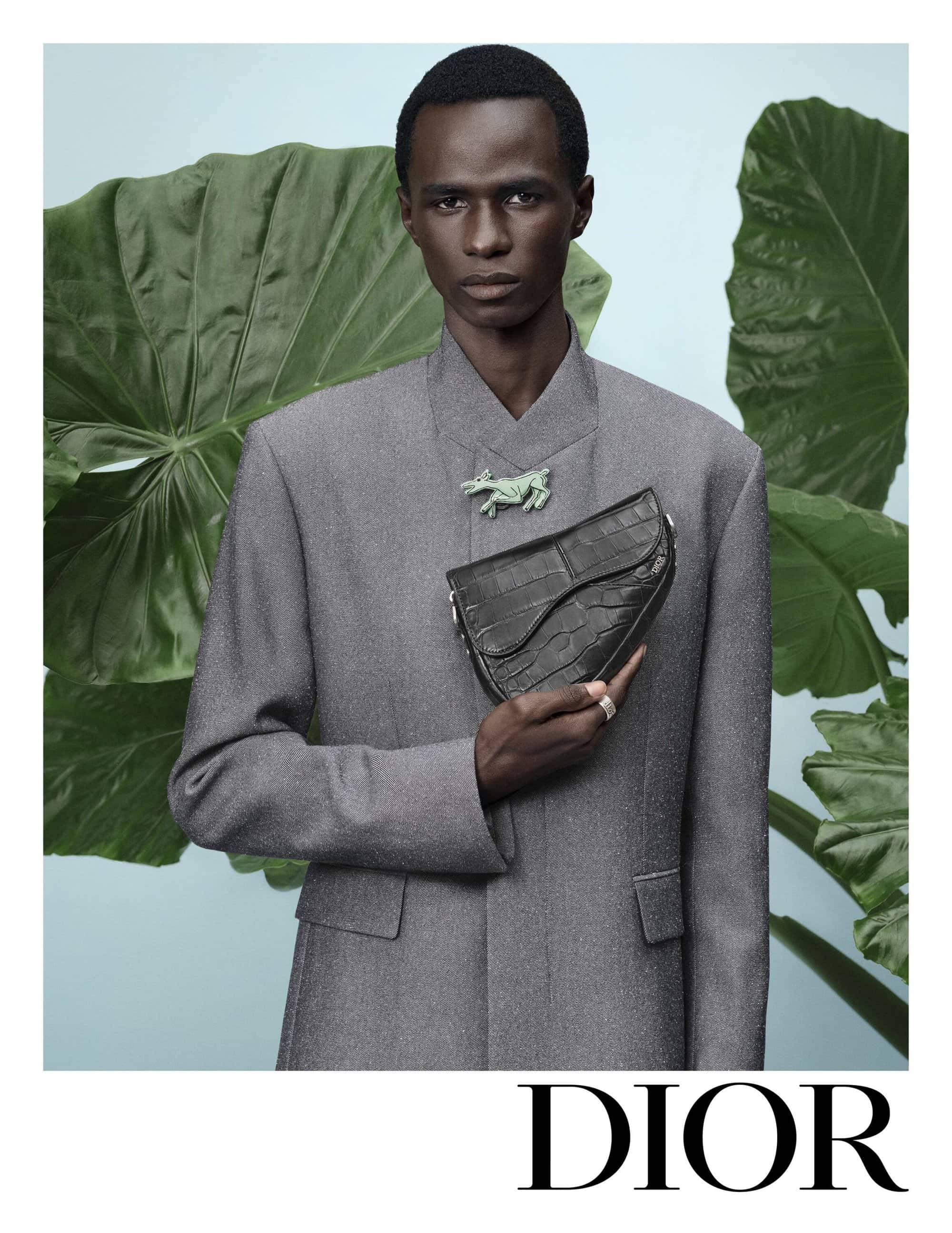 Black model wearing a grey jacket holding a black saddle bag against large green leaves