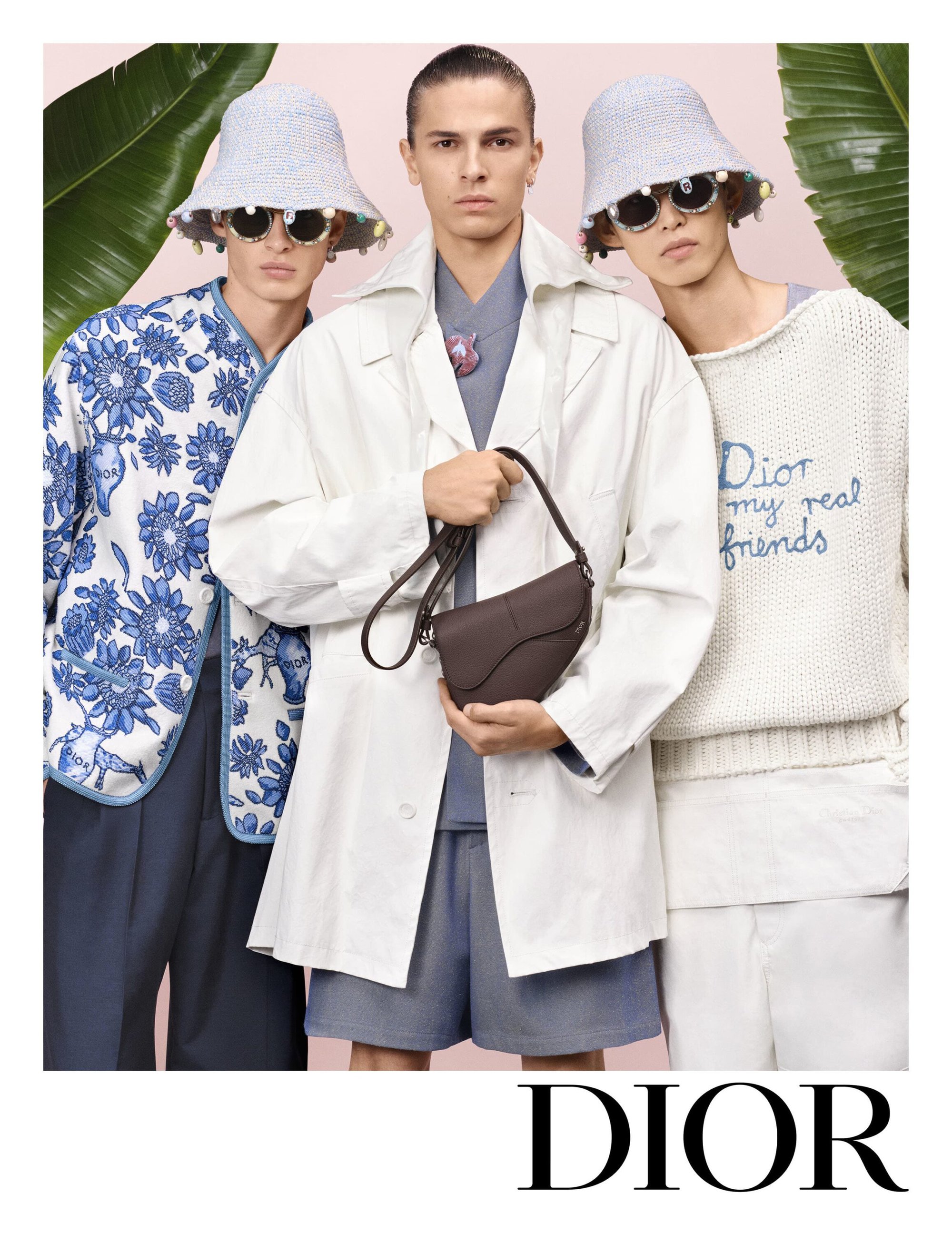 Three male models posing together against a soft pink background framed by green banana leaves, wearing Dior Spring 2025 menswear