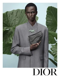 Black model wearing a grey jacket holding a black saddle bag against large green leaves