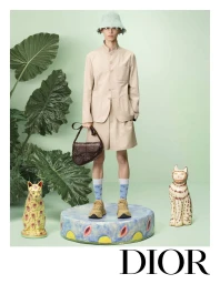 Model standing on a decorated round plinth wearing a beige jacket, shorts, hat, and holding a brown handbag against a pale green backdrop.