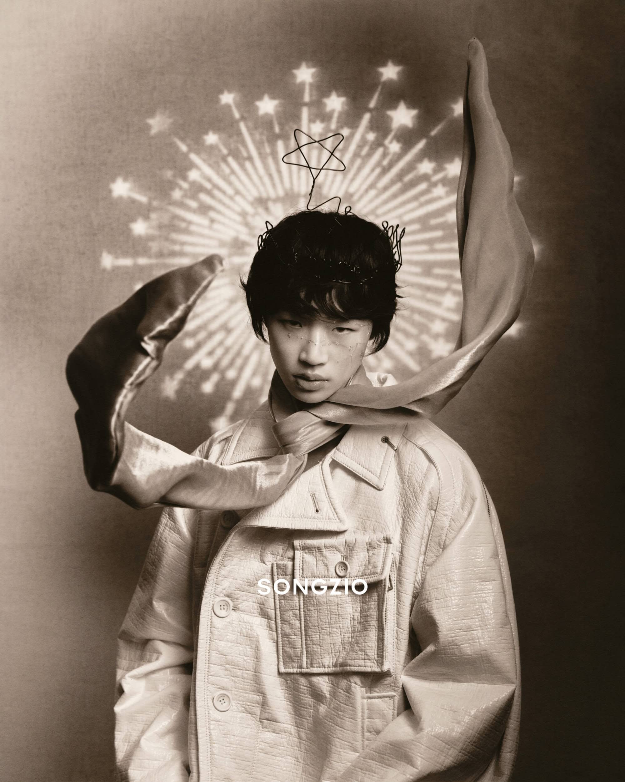 Portrait of young man wearing a light textured jacket and star crown with a flowing scarf in front of a projected starburst background