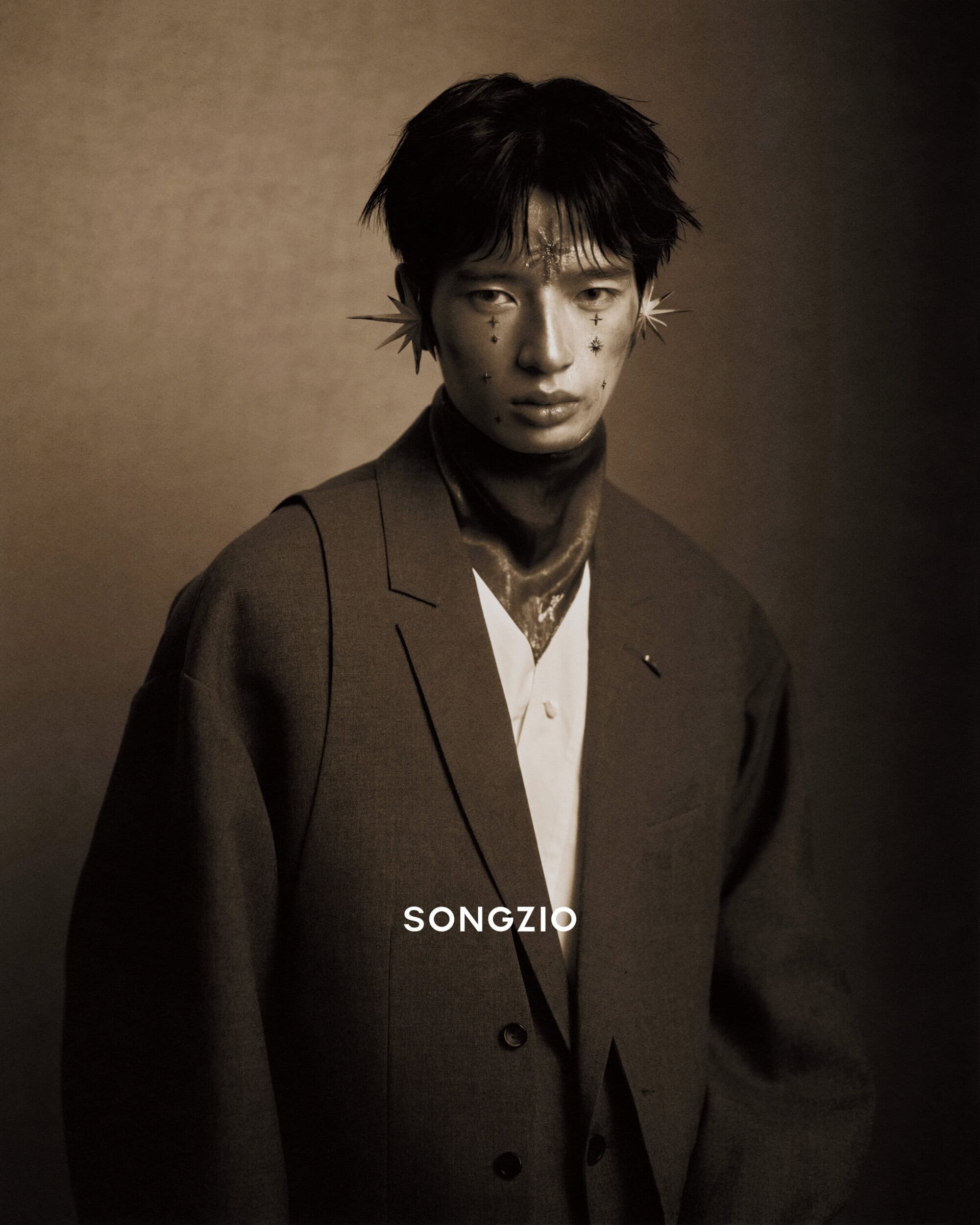 East Asian male model wearing a dark grey blazer and star-shaped ear cuffs against a dark textured studio backdrop.