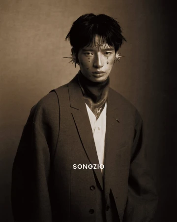 East Asian male model wearing a dark grey blazer and star-shaped ear cuffs against a dark textured studio backdrop.