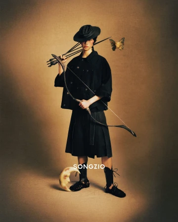 Model in black hat and dark attire holding a bow and arrows against a textured brown gradient background with a floating butterfly and crescent moon