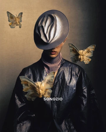 Model wearing a grey hat and dark jacket surrounded by floating butterfly sculptures with human faces for Songzio