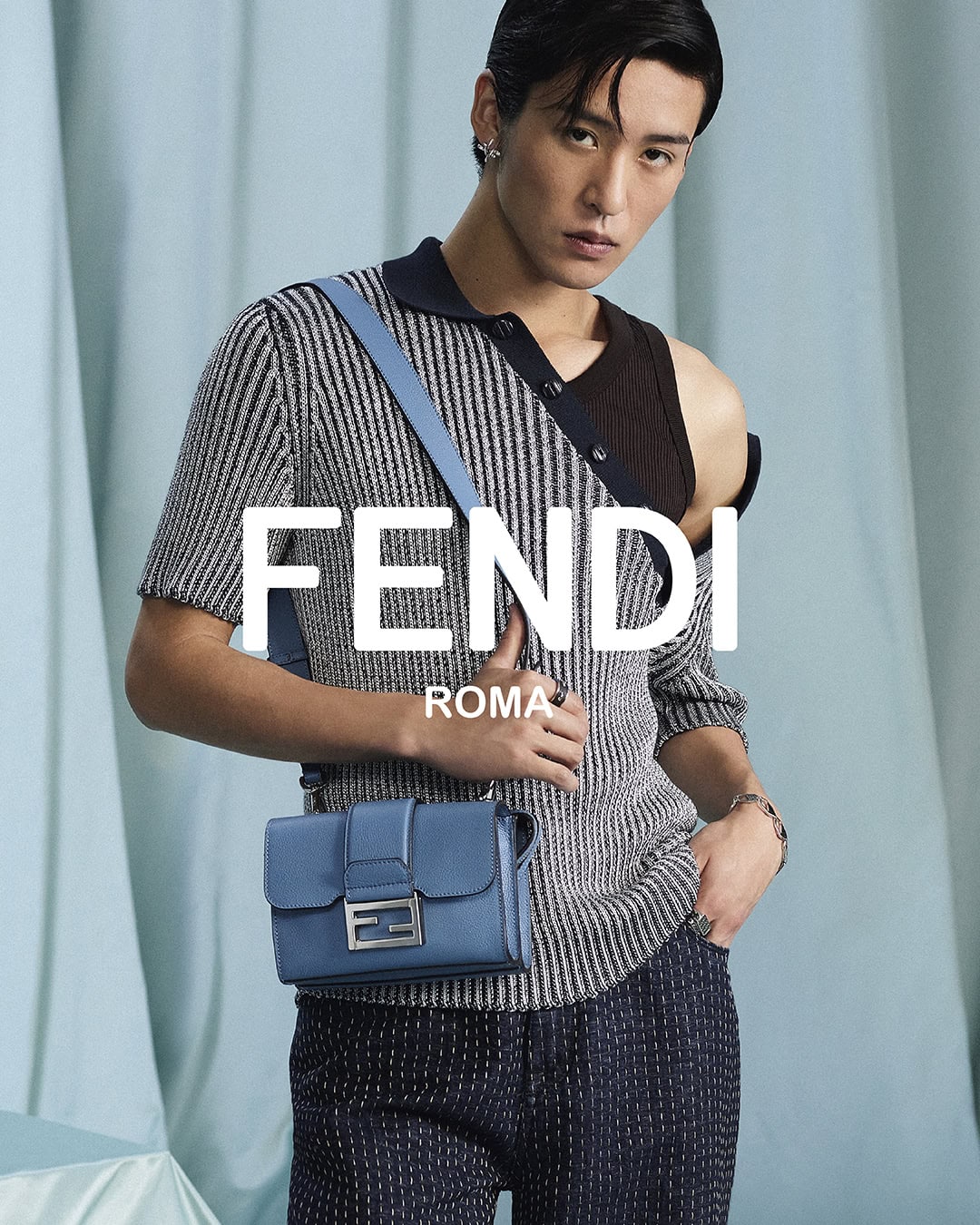 Model Ren Meguro wearing a grey patterned top and holding a blue Fendi crossbody bag against a draped teal curtain backdrop.