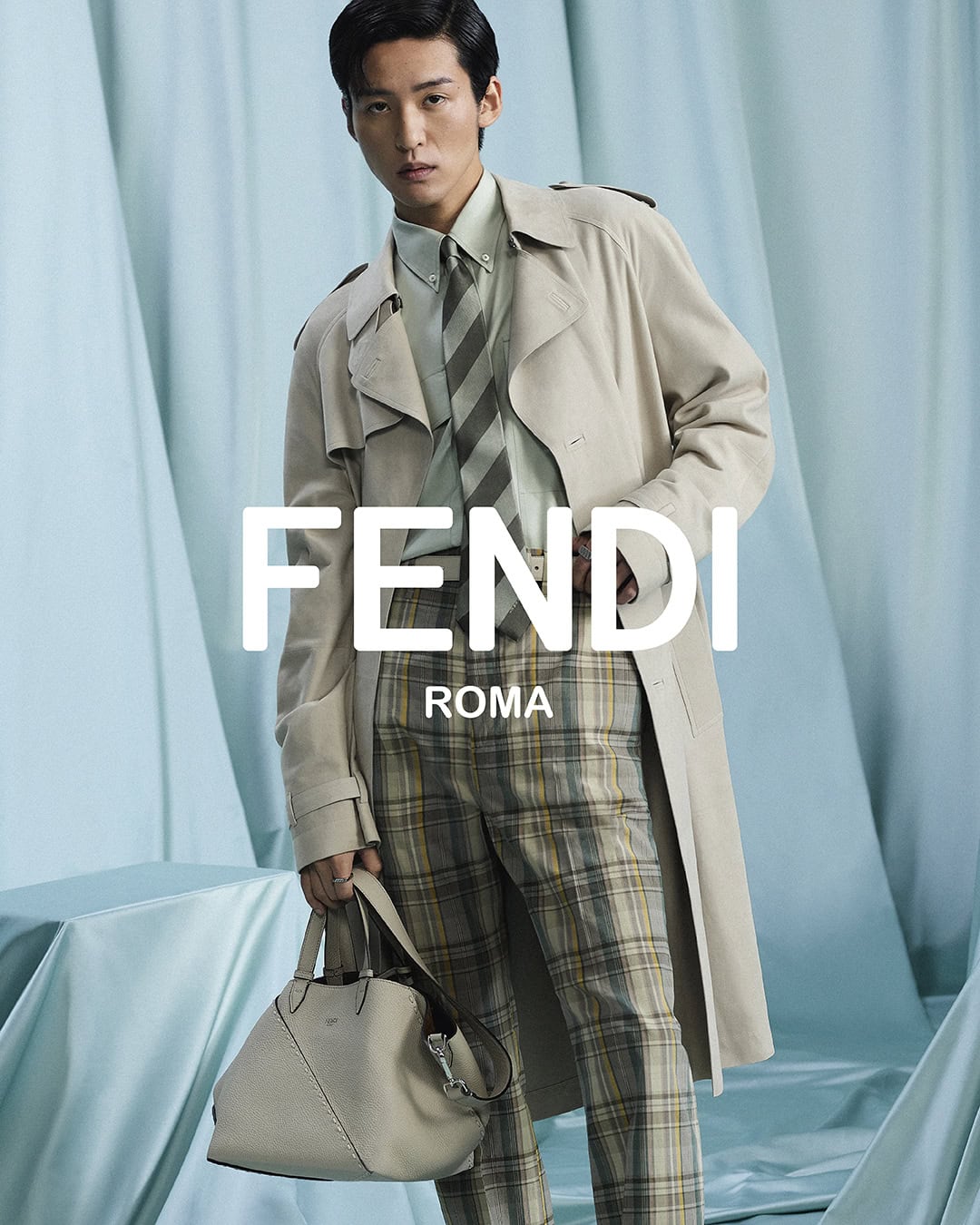 Model Ren Meguro wearing a beige trench coat and plaid trousers holding a handbag against a draped pale blue backdrop.