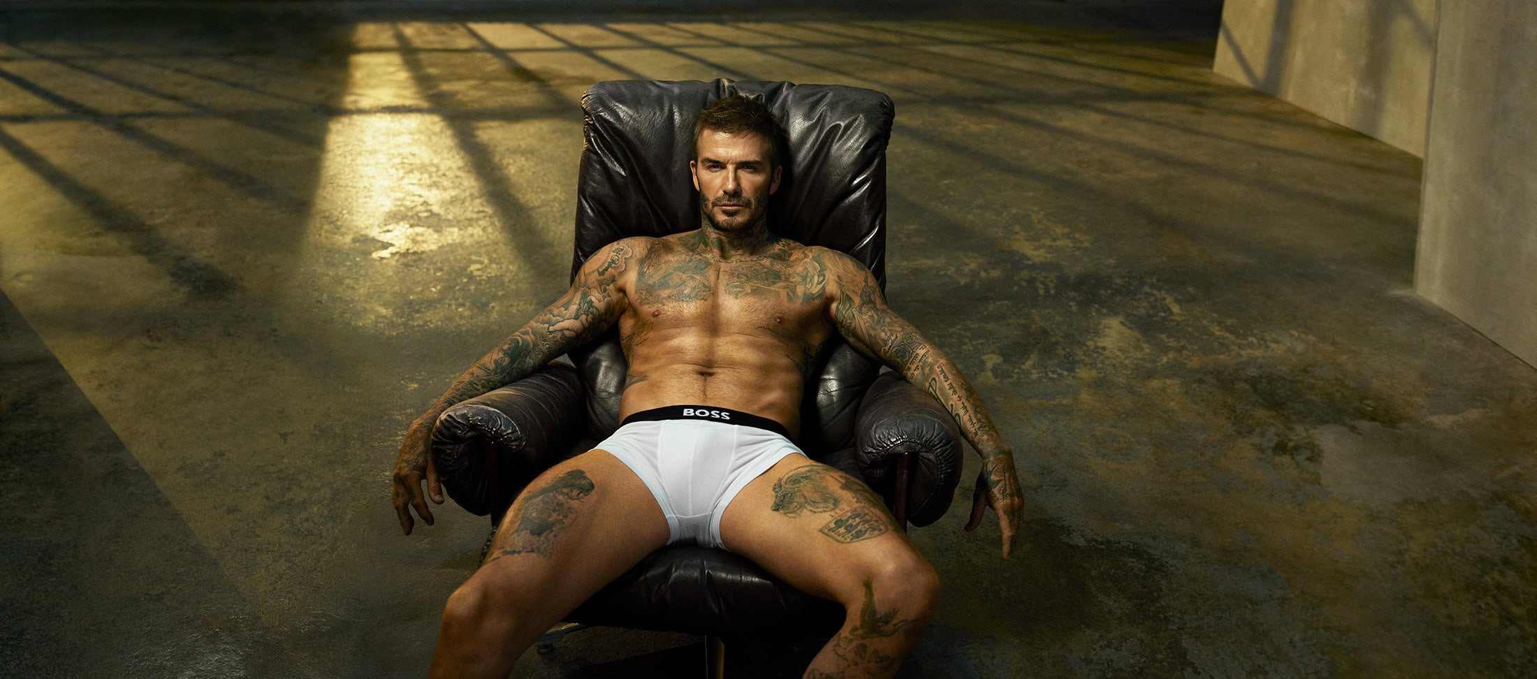 David Beckham wearing white BOSS briefs seated in a black leather armchair on a concrete studio floor.